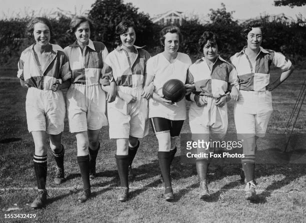 Line of six women footballers, five wearing shirts with a quartered design, and one player carrying a football wearing a white shirt, posing...
