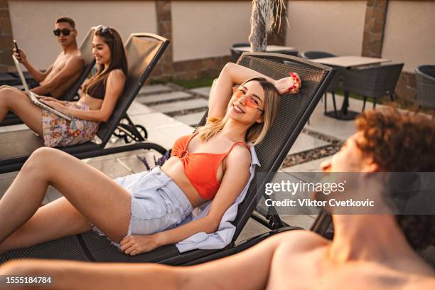 small group of friends laying down on sunbeds by the swimming pool - zonnebank stockfoto's en -beelden