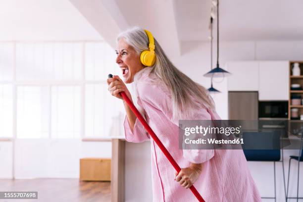 carefree woman with yellow headphones listening to music holding broom at home - escoba fotografías e imágenes de stock