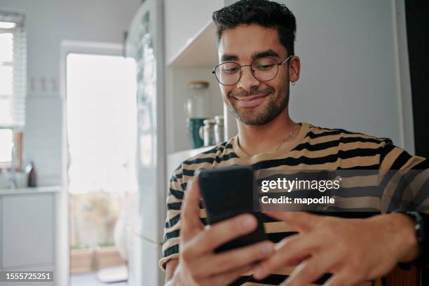 jeune homme souriant assis dans sa cuisine et faisant défiler son téléphone intelligent - rencontre sur internet photos et images de collection
