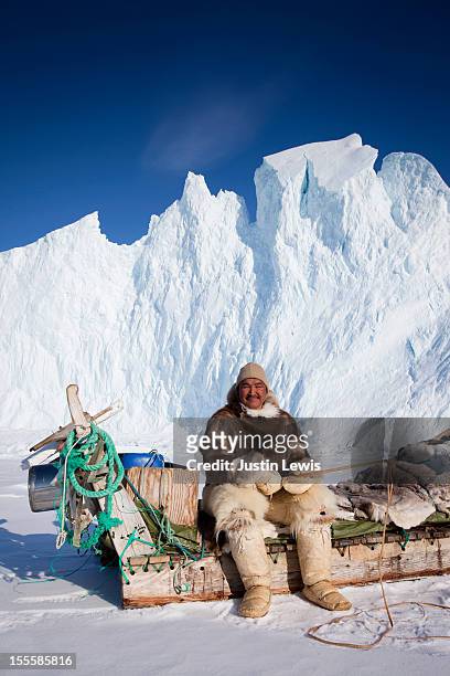 inuit male in fur sits on sled in front of iceberg - inuit people stock-fotos und bilder