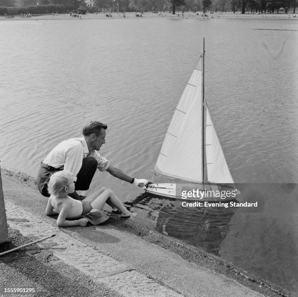 Man launches a model sailing boat into a pond while a boy next to him reclines with his feet in the water, London, August 4th 1959.