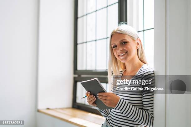 portrait of young woman student with long blonde hair making notes in notebook near windowsill - business student stock-fotos und bilder