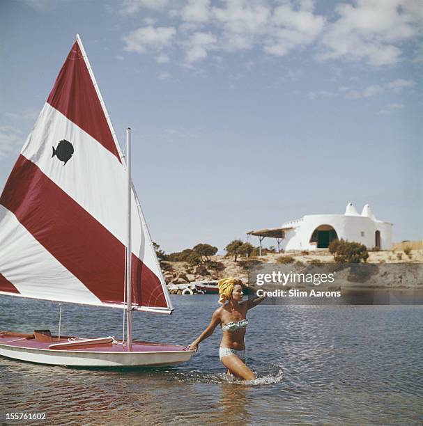 French fashion model Bettina Graziani wading in the shallow waters of Costa Smeralda, pulling a small yacht behind her, in Sardinia, Italy, 1964.