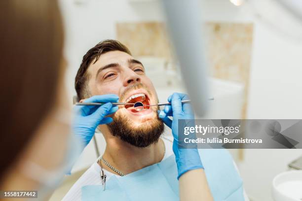 male patient sits calmly in dentist chair while dentist uses dental mirror to inspect teeth - toothache stock-fotos und bilder