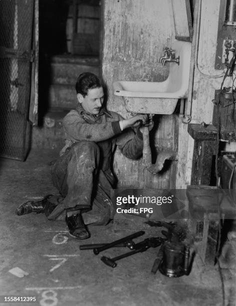 Plumber wearing overalls sits on the floor as he fits a drain pipe to the underside of a sink, his tools arranged on the floor before him, United...