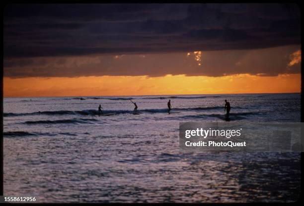 View of the silhouettes of four surfers on the water at sunset, Hawaii, October 1965.