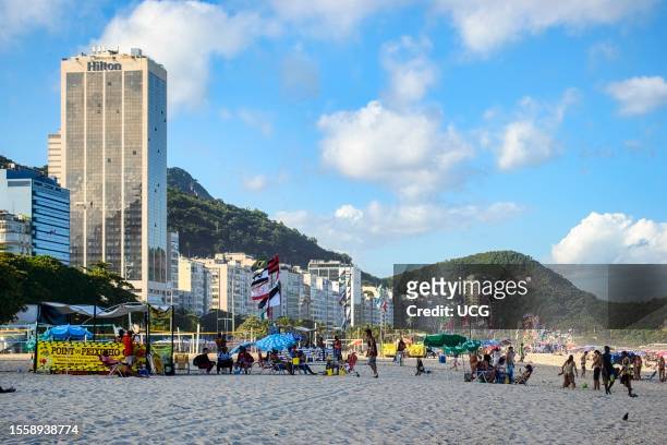 Rio de Janeiro, Brazil, Groups of people populate Copacabana beach on a sunny day. A cityscape of tall buildings, one named Hilton, decorates the...