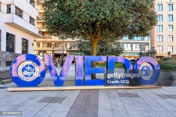 Oviedo, Asturias, Spain, A letter sculpture spelling "Oviedo" surrounded by tiled flooring in a downtown district. Trees, city buildings, and...