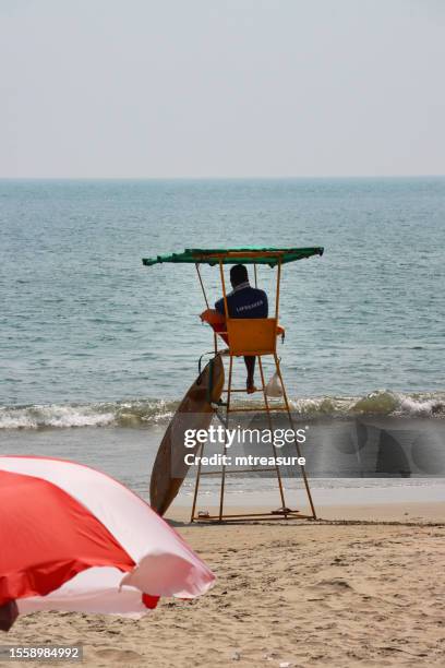 close-up image of beach lifeguard sat in lifeguard observation chair, paddle board, watching sea and coastline of palolem beach, goa, india - surf rescue stock pictures, royalty-free photos & images