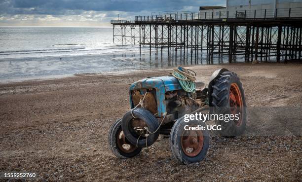 Vintage and rusted tractor used by a fisherman to bring in his boat, lies parked on the shingle beach in front of Bognor Regis Pier, in West Sussex,...