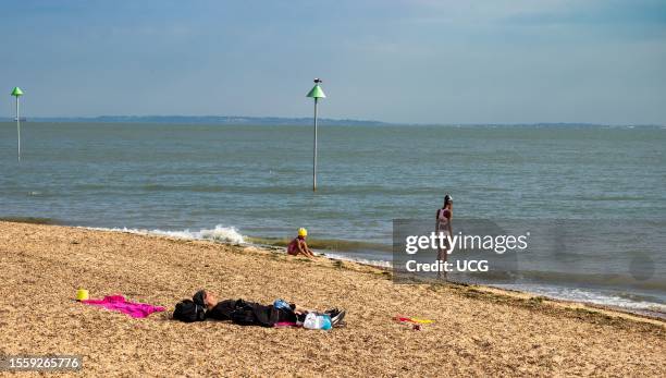Young black girl stands at the edge of the sea, while her younger sister plays nearby and a women sleeps in the sun on Southend-on-Sea beach, Essex,...
