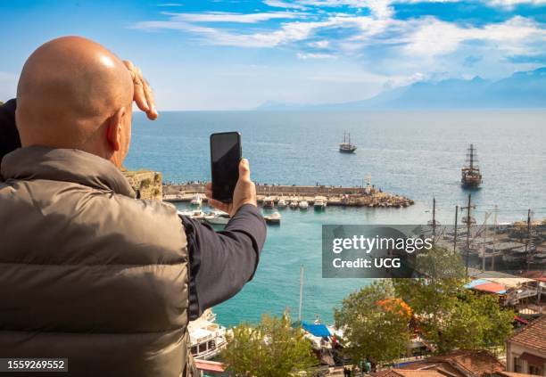 Man with a bald head takes a photograph of tourist boats returning to the ancient Harbor at Antalya in Turkey. The bustling harbor is busy with...