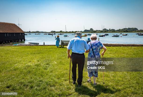 An elderly couple in their 90s walk across a grassy area in Bosham next to Chichester Harbor, UK.