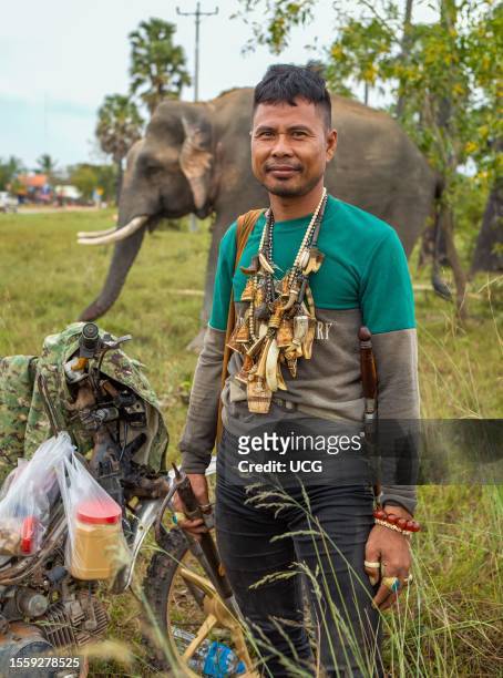 Cambodian Mahout adorned with Buddhist amulets to ward off danger stands close to his elephant on scrub land in Phumi Khna, Siem Reap Province,...
