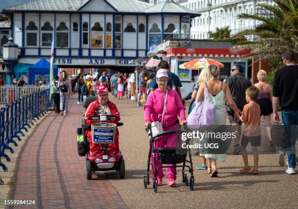 Disabled couple use a mobility scooter and a rollator on the promenade next to Eastbourne Pier, East Sussex, UK.