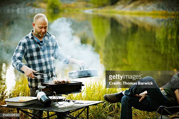 man barbecuing on grill near mountain lake - mann lässig gras sitzen stock-fotos und bilder