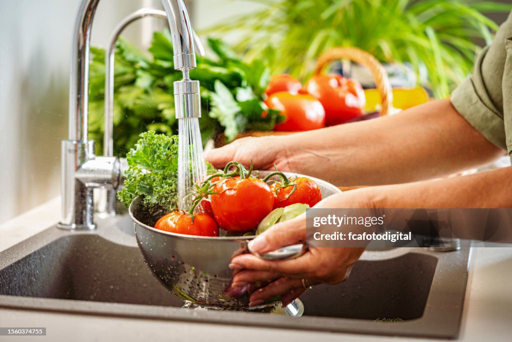 Mujer sosteniendo un colador lleno de verduras frescas bajo agua corriente en el fregadero de la cocina