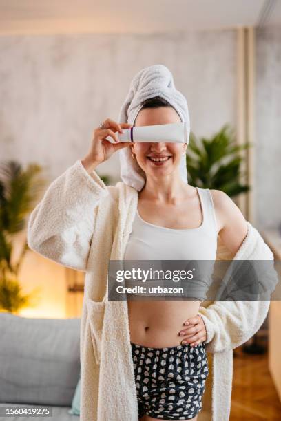 young woman holding up a blank moisturizer product in the living room - robe tube stock pictures, royalty-free photos & images