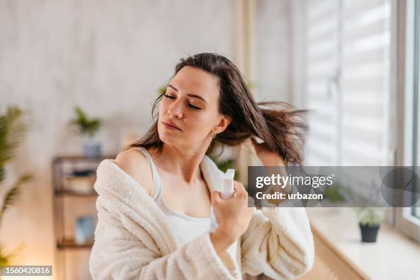 mujer joven rociando un producto para el cabello en su cabello en la sala de estar - cabello humano fotografías e imágenes de stock