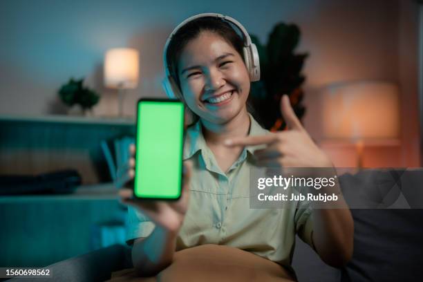 smiling asian woman holding her phone with a green screen in her room at night. - green screen stock pictures, royalty-free photos & images