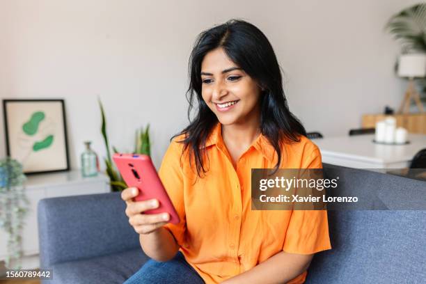 happy young indian female looking at smartphone screen while sitting on comfortable couch at home. technology and social media concept. - indian ladies stock pictures, royalty-free photos & images