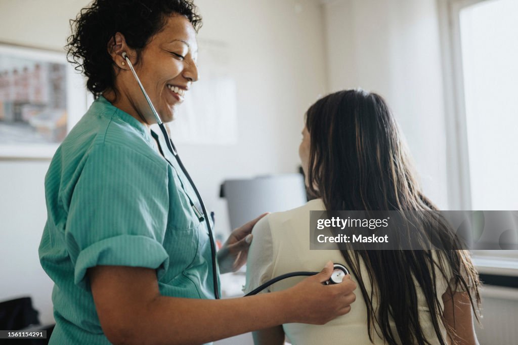 Side view of smiling mature female doctor examining young woman with stethoscope in clinic