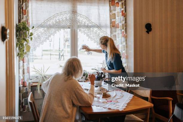 senior woman looking at female caregiver cleaning window glass at home - poetshulp stockfoto's en -beelden