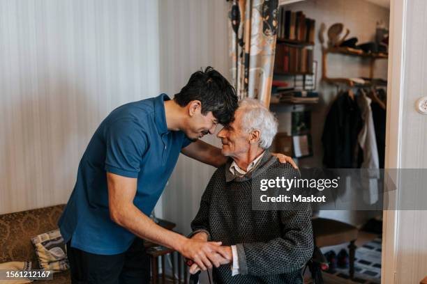 smiling male care assistant touching forehead with senior man at home - asistencia de la comunidad fotografías e imágenes de stock