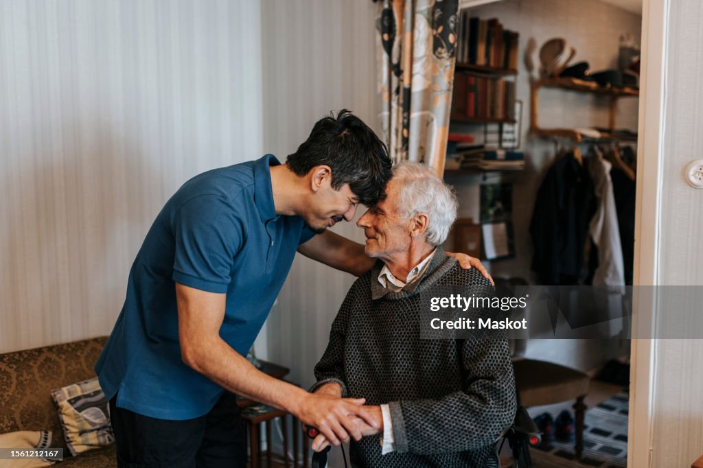 Smiling male care assistant touching forehead with senior man at home