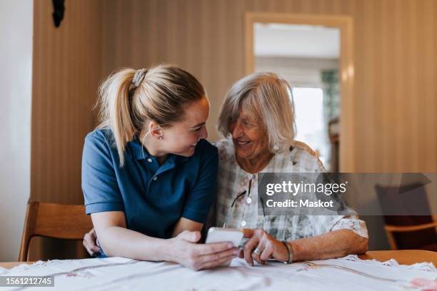 smiling female care assistant sharing smart phone with senior woman while sitting at dining table in home - asistencia de la comunidad fotografías e imágenes de stock