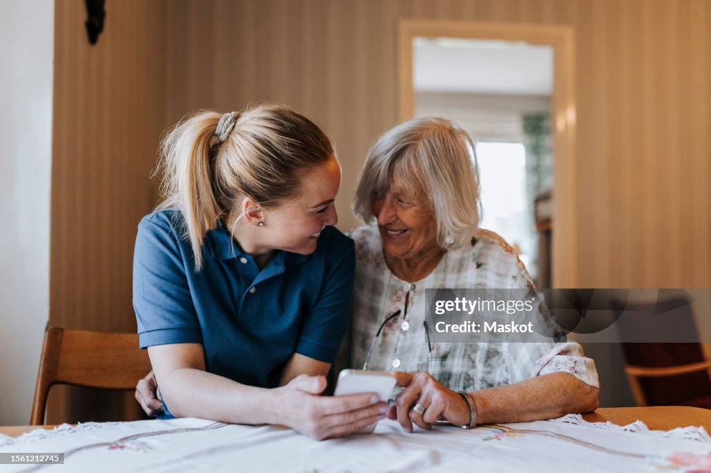 Smiling female care assistant sharing smart phone with senior woman while sitting at dining table in home