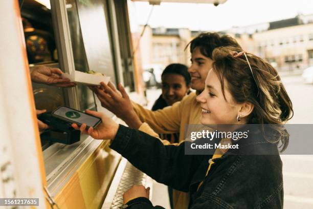 smiling young woman doing online payment through smart phone while buying food from concession stand - arab teen photos et images de collection