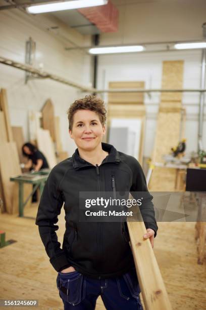 portrait of confident mature female carpenter carrying plank while standing in workshop - wood worker posing stock-fotos und bilder