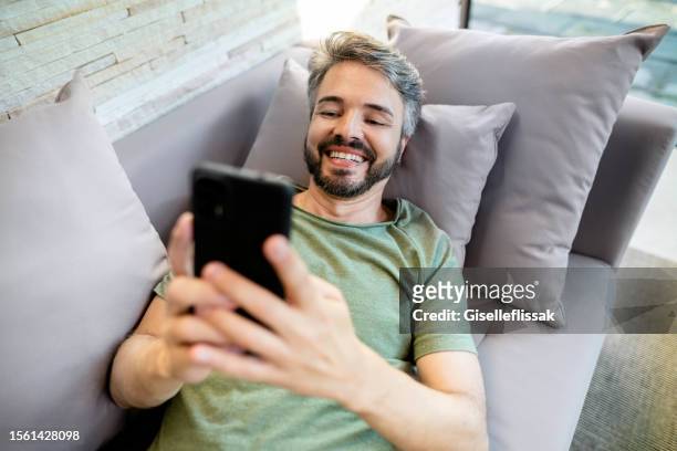 happy mid adult man text messaging on his phone on a sofa at home - sending sms stock pictures, royalty-free photos & images