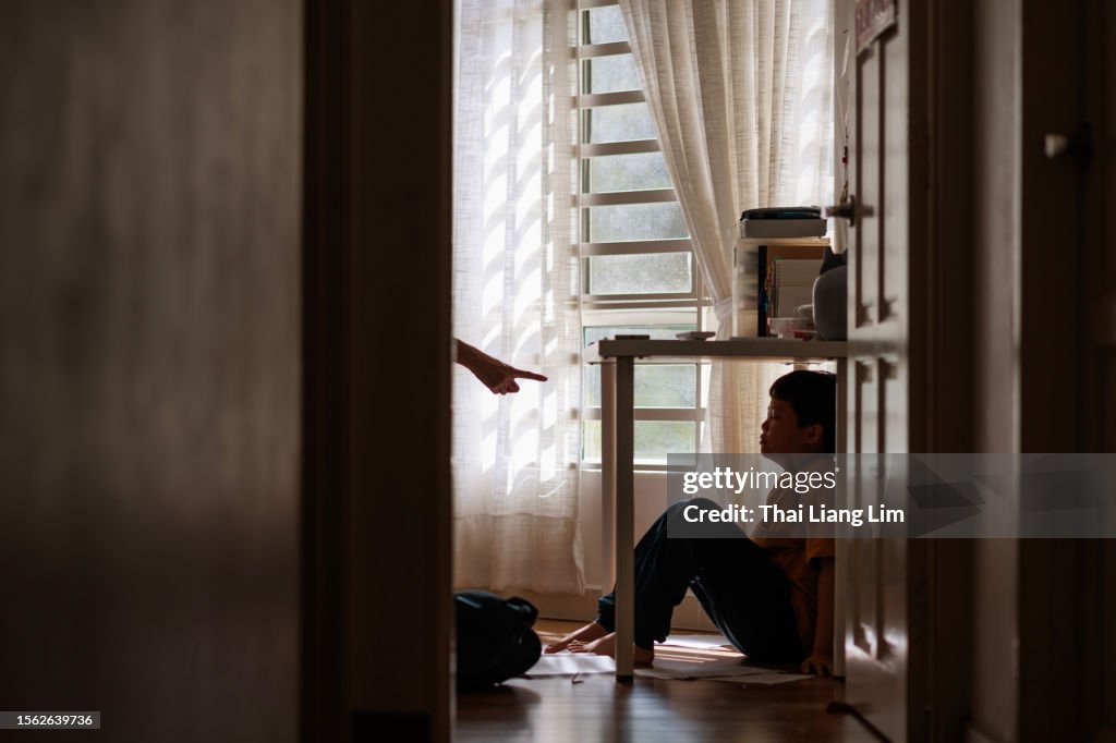 A little Asian boy with a sad and stress expression hides in the shadows under a table at home, while being scolded by his mother