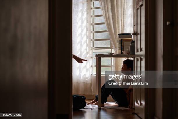 a little asian boy with a sad and stress expression hides in the shadows under a table at home, while being scolded by his mother - abuse 個照片及圖片檔