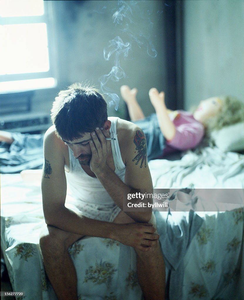 Man Smoking on Bed with Blow Up Doll in Background