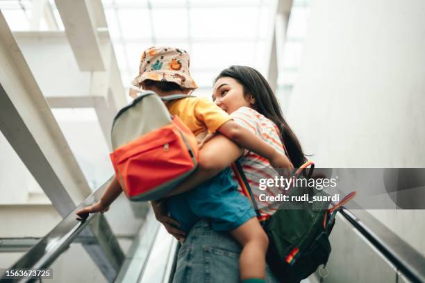 mutter und sohn tragen rucksack, während sie auf einer rolltreppe fahren und zur schule pendeln - toddler at airport stock-fotos und bilder