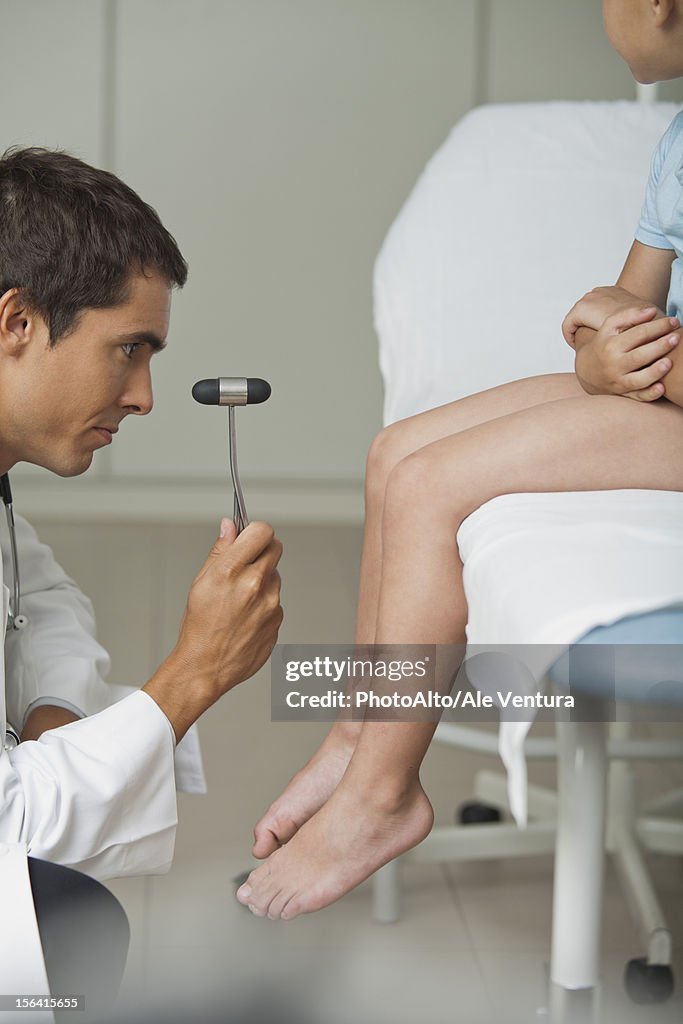 Doctor checking boy's reflexes with reflex hammer, side view, cropped