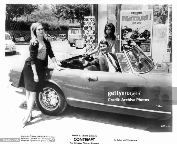 Brigitte Bardot leaning against car as Jack Palance looks over to her in a scene from the film 'Contempt', 1963.