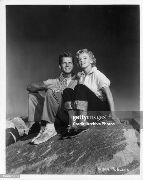 Keith Andes and Marilyn Monroe sitting on rocks together in publicity portrait for the film 'Clash By Night', 1952.