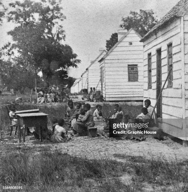 The children of enslaved people sit outside their quarters, one of a row of huts extending into the distance, on a plantation in Charleston, South...
