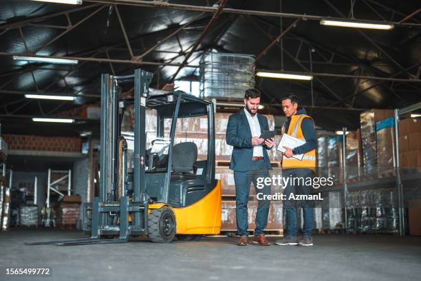 dos hombres multiétnicos parados junto a una carretilla elevadora en un almacén - carretilla elevadora fotografías e imágenes de stock