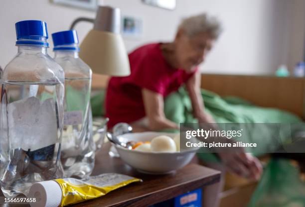 July 2023, Berlin: Water bottles, a bowl of food and a cream lie on a bedside table by the bed of a senior woman in need of care, taken during a...