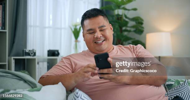 chubby man using smartphone to relaxing on sofa in living room - obesity stock pictures, royalty-free photos & images