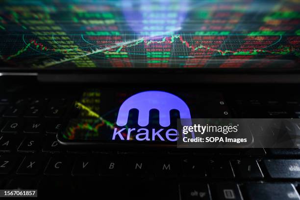 In this photo illustration a Kraken logo seen displayed on a smartphone with stock market percentages on a laptop screen.