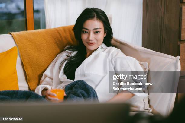 young asian woman lying on the sofa bed enjoying the weekend, watching movie on tv and eating a food at home in the evening, relaxation time indoor a house, young woman watching news on television - asian watching tv stock pictures, royalty-free photos & images