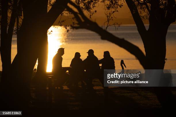 August 2023, Australia, The Entrance: People sitting next to a pelican at dusk at Tuggerah Lake. Photo: Sebastian Gollnow/dpa