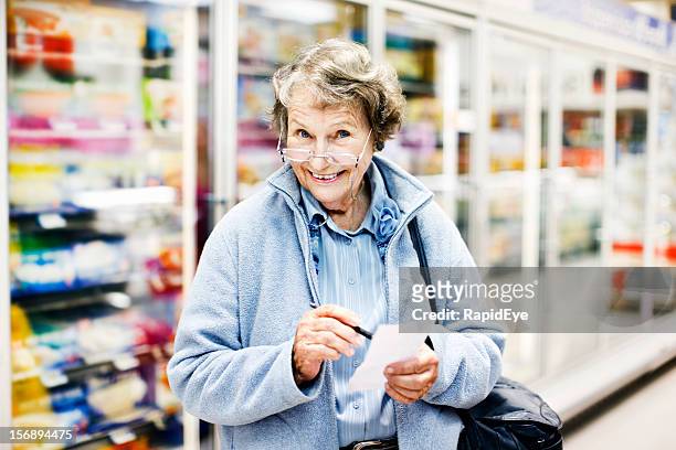 senior woman in supermarket checks shopping list, smiles delightedly - senior women shopping stock pictures, royalty-free photos & images